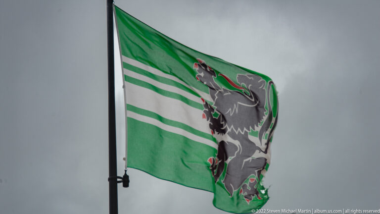 Green-and-white striped flag with Ghent's black lion emblem blowing strongly against a gray sky.