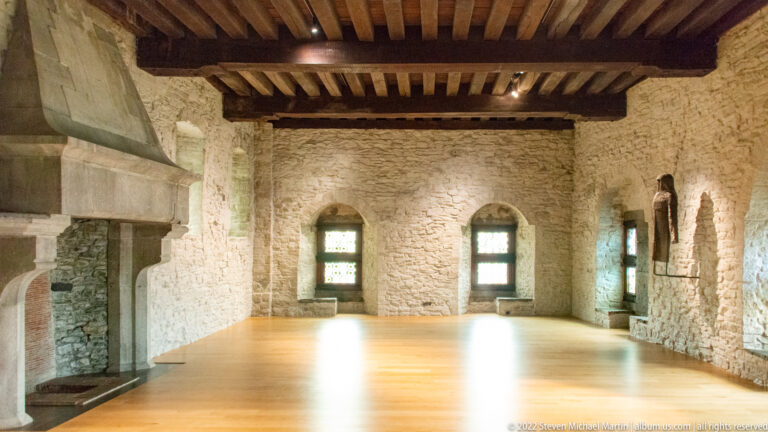 Large castle room with rough stone walls, timber ceiling beams, wooden floor, recessed windows, and a stone fireplace.