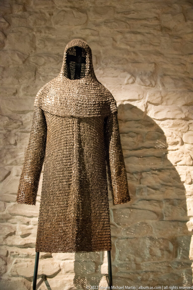 Short-sleeved chain-mail shirt displayed on a stand against a rough stone wall.