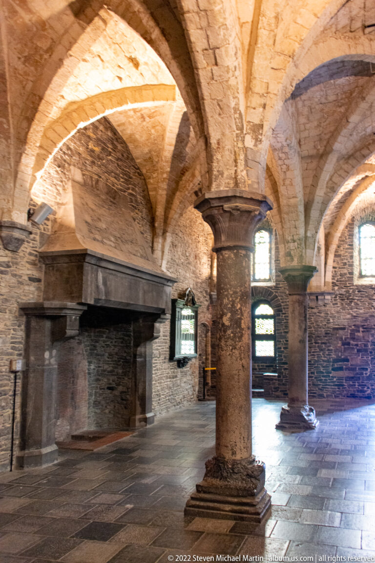 Stone-vaulted castle room with a central column, fireplace, small windows, and rough masonry walls.