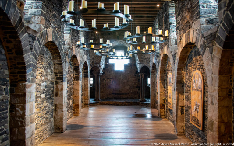 Long stone gallery with repeated arches, timber ceiling, circular chandeliers, and interpretive panels inside Gravensteen.