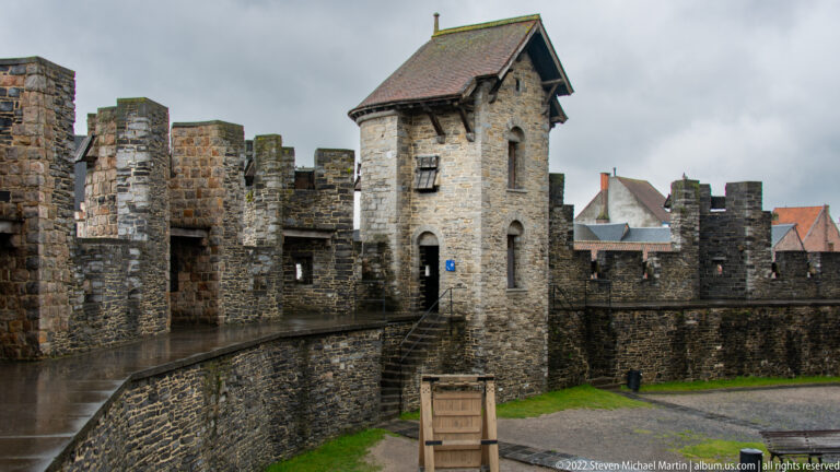 Roofed stone tower connected to crenellated castle walls by a narrow elevated walkway inside Gravensteen.