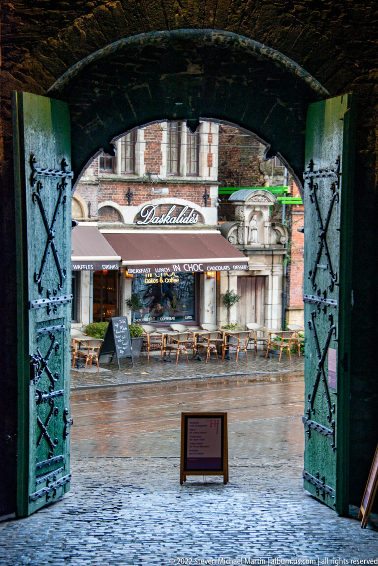 Open heavy castle doors framing a wet Ghent street and café beyond from inside Gravensteen.