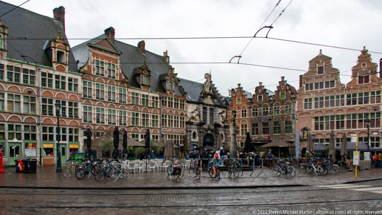 Historic gabled façades around Sint-Veerleplein with bicycles, café seating, tram tracks, and wet pavement.