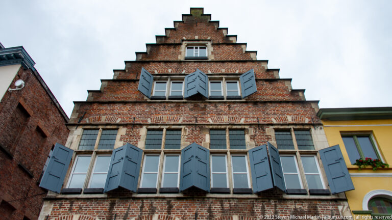 Brick stepped-gable house with blue-gray shutters, multi-pane windows, and pale stone trim viewed from below.