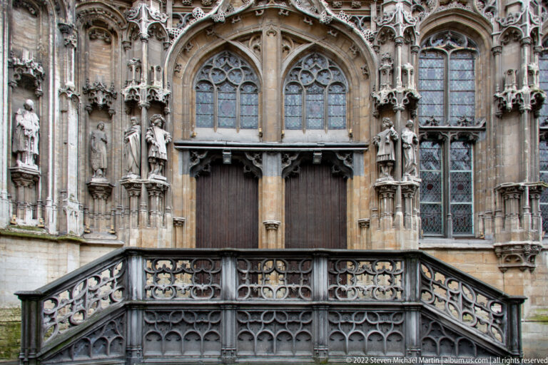 Highly ornate Gothic church façade with pointed windows, sculpted figures, tracery, wooden doors, and carved stone balustrade.