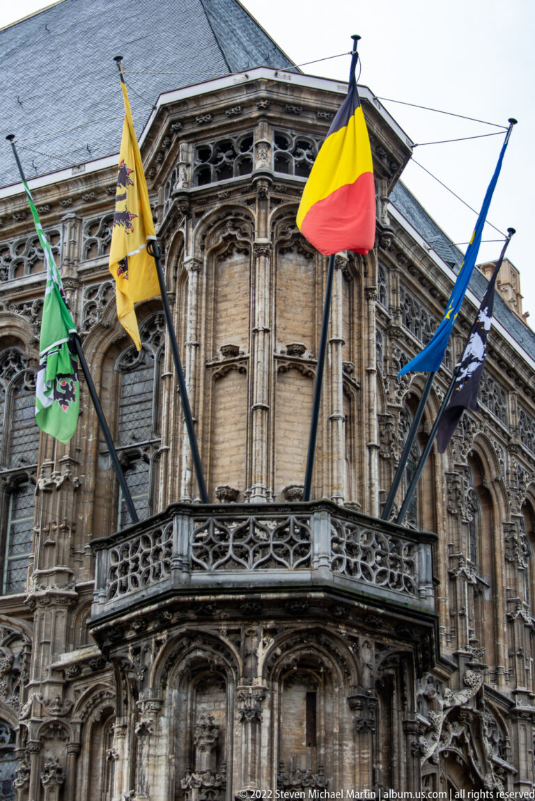 Belgian tricolor and several heraldic flags flying from poles above a carved balcony on an ornate stone façade.
