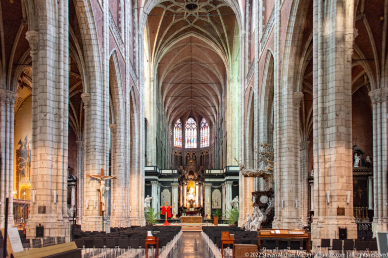 Central nave of Saint Bavo's Cathedral with tall stone piers, ribbed vaults, stained glass, and illuminated altar.