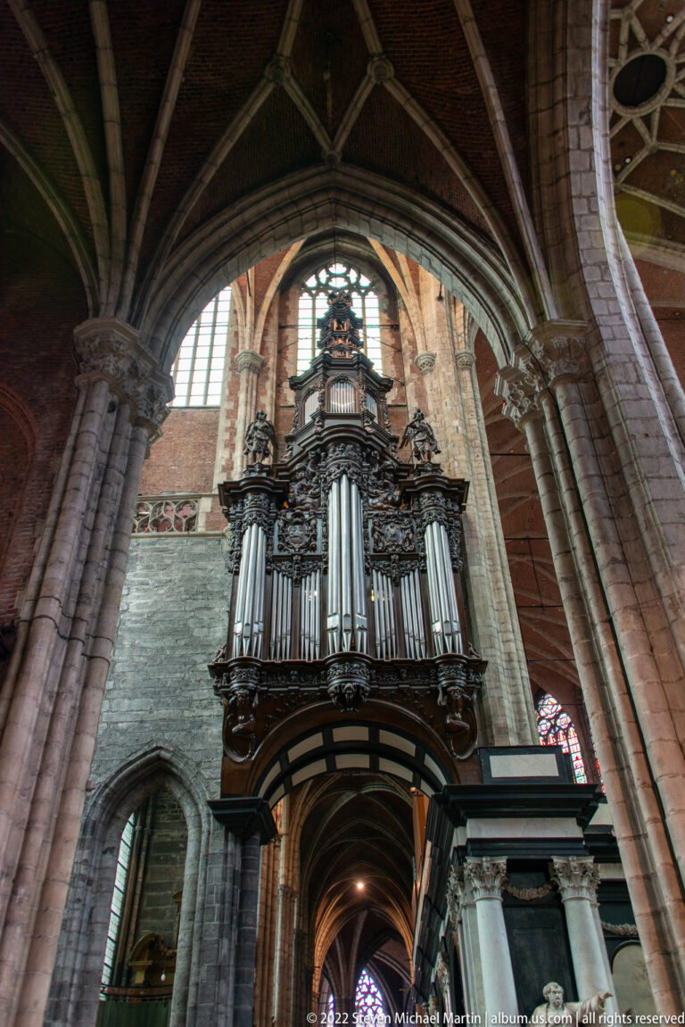Large dark-cased pipe organ with silver pipes mounted high among Gothic arches and clustered stone columns.