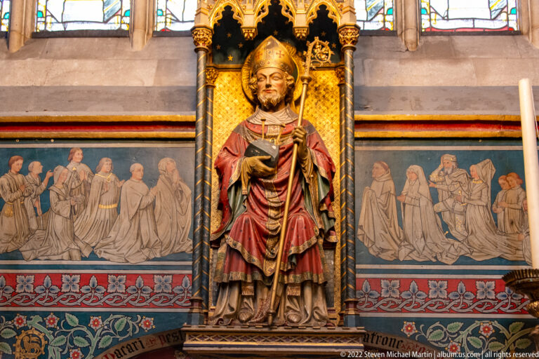 Polychrome bishop saint in mitre and red vestments beneath a gilded canopy, with painted kneeling figures behind.