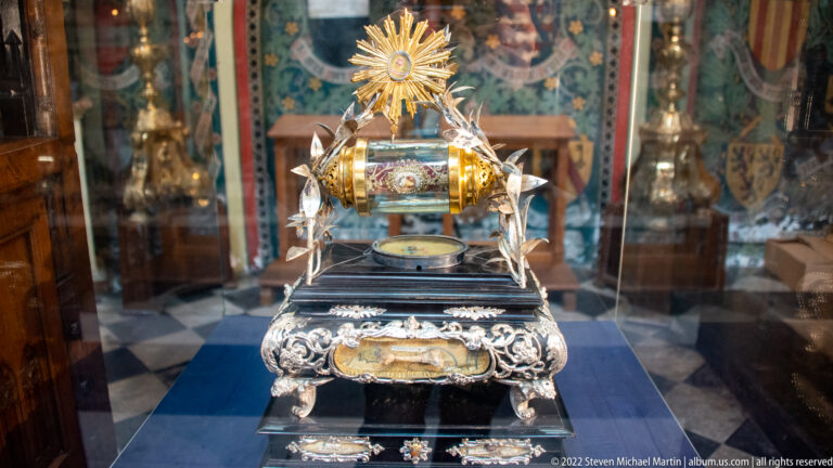 Ornate silver-and-gilt devotional vessel with radiant sunburst, winged supports, decorated base, and central receptacle in a glass case.