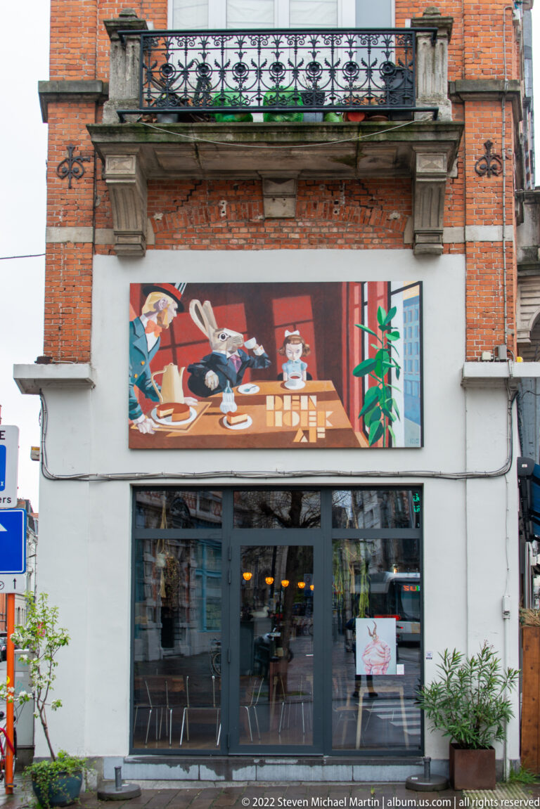Gray storefront with broad glass doors and a colorful mural of stylized figures, dishes, and plants above the entrance.