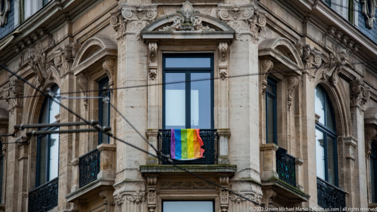 Rainbow flag hanging from a small balcony on an ornate historic stone façade with carved window surrounds.
