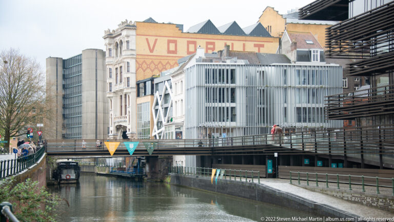 Urban canal with a pedestrian bridge, modern metal-and-glass buildings, and older rooftops in Ghent.