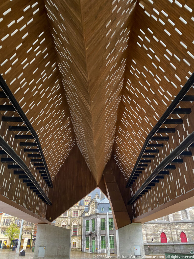 Angular wooden canopy with perforated slats and dark steel framing, viewed upward above historic Ghent façades.