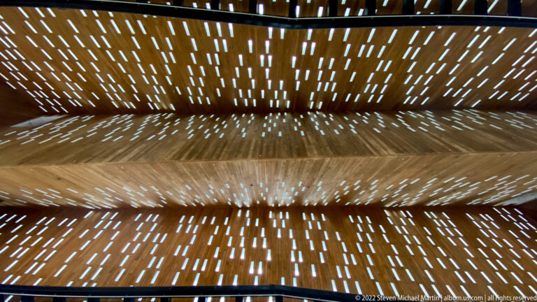 Close upward view of slatted wooden roof planes pierced by many narrow openings of daylight.