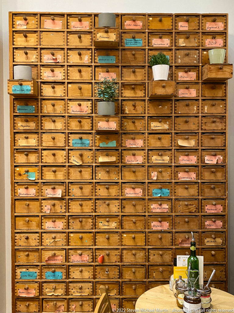 Large wall of many small labeled wooden drawers with pastel labels, plants, and objects in a hospitality interior.