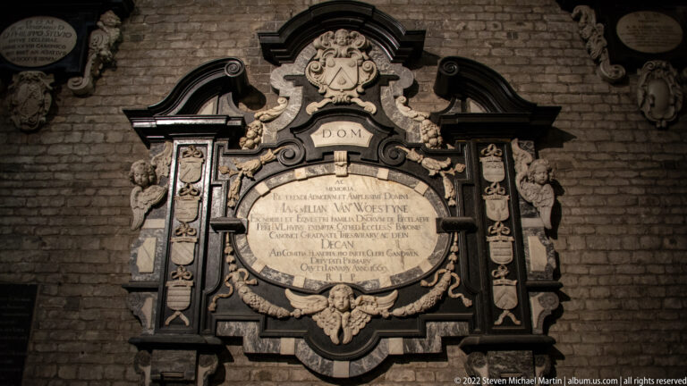 Ornate black-and-white wall memorial with Latin inscription, scrollwork, skulls, and cherubic ornament on brick.