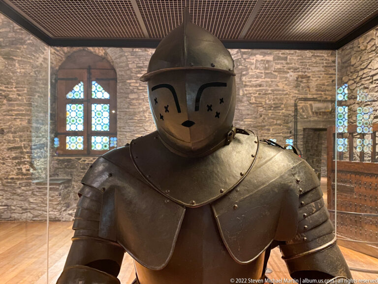 Full dark steel suit of plate armor with broad pauldrons and closed helmet displayed in a museum case.