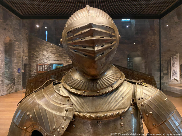 Polished closed helmet with horizontal visor slits, fluted neck defense, rivets, and upper breastplate.