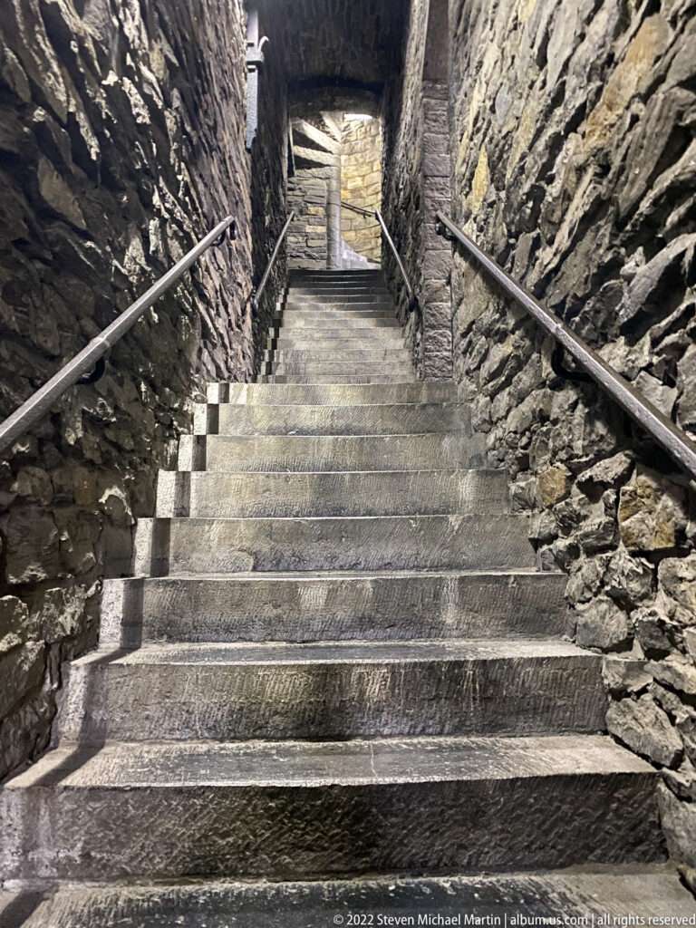 Steep stone stairs rising between rough masonry walls with metal handrails toward a lit upper landing.