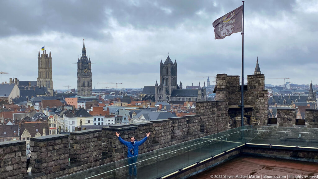 Visitor with raised arms on Gravensteen rooftop terrace overlooking Ghent's historic towers and dense city skyline.