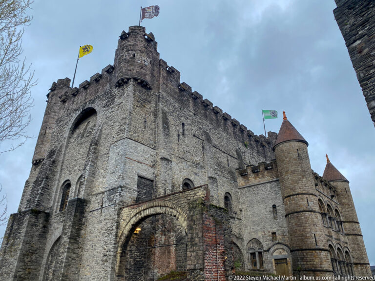 Low-angle view of Gravensteen's tall stone keep, crenellations, flags, and round conical-roofed corner towers.