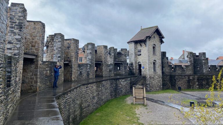 Wet rampart walkway inside Gravensteen with crenellated walls, stone towers, a roofed watchtower, and one visitor.