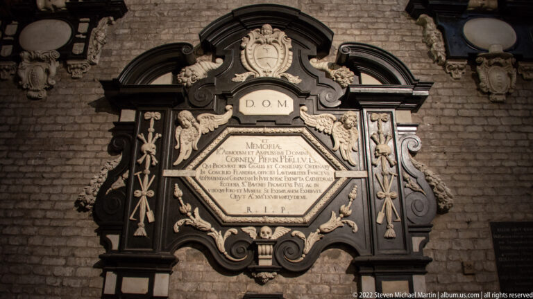 Dark ornate funerary wall monument with pale Latin cartouche, scrolls, columns, and skull motifs on brick.