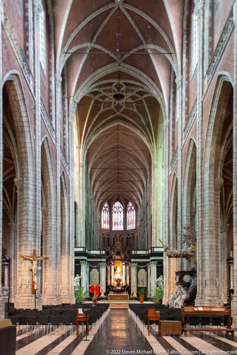 Wide symmetrical view of Saint Bavo's Cathedral nave with pale stone piers, reddish ribbed vaults, pews, and altar.