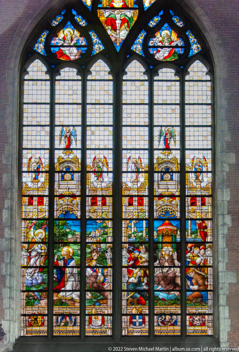 Large multi-light Gothic stained-glass window with narrative scenes, heraldic shields, and clear-glass panels.