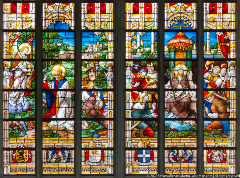 Close view of colorful stained-glass scenes with groups of figures, architectural details, and heraldic shields.