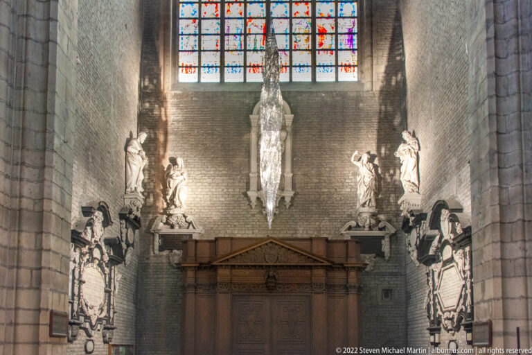 Brick cathedral bay with wall memorials, statues, stained glass, and a pale suspended sculpture above a wooden enclosure.