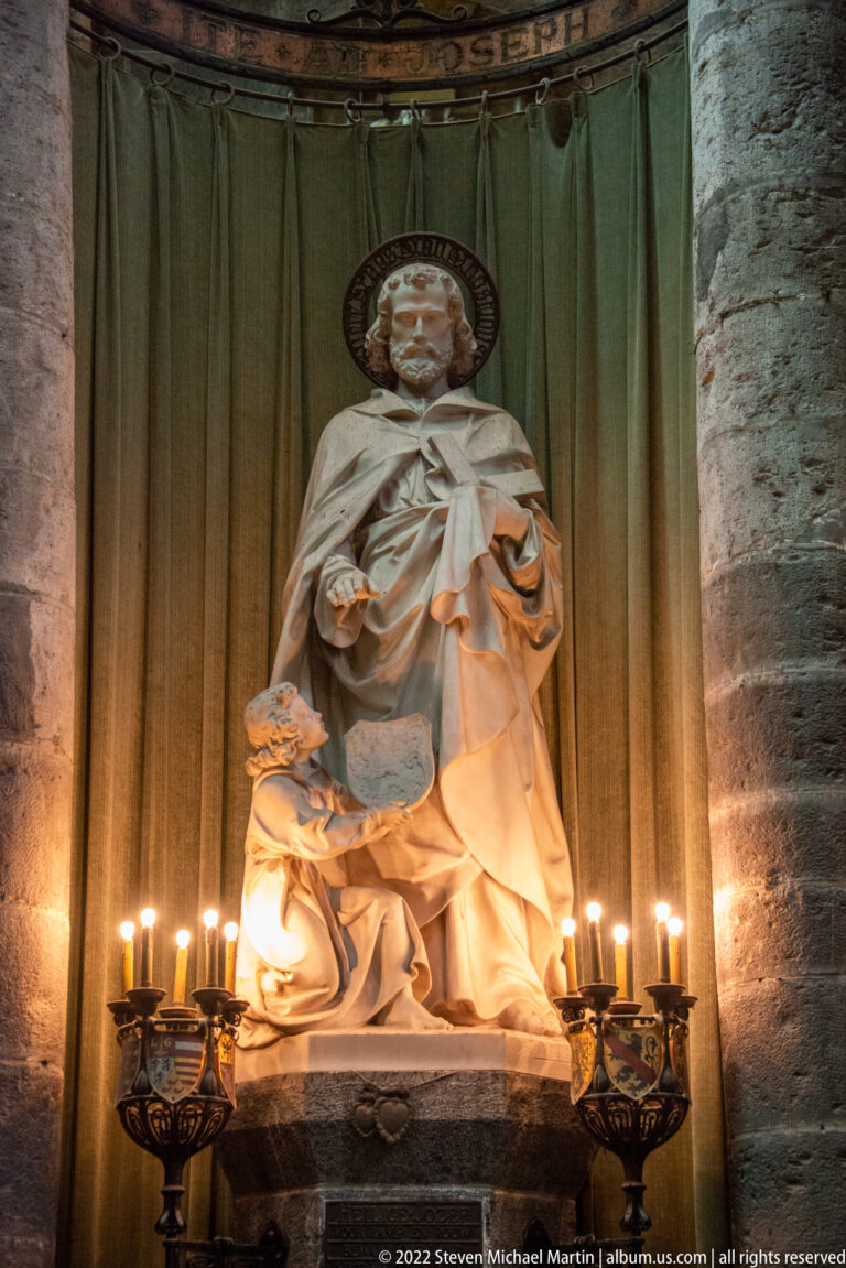 Seated haloed saint in pale stone holding a book, with a smaller figure beside him and candles on both sides.