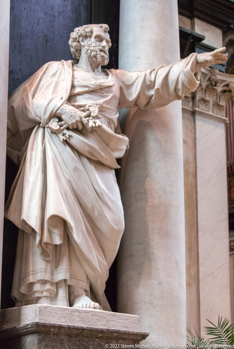 Large pale stone statue of a bearded robed saint pointing outward beside a classical column.