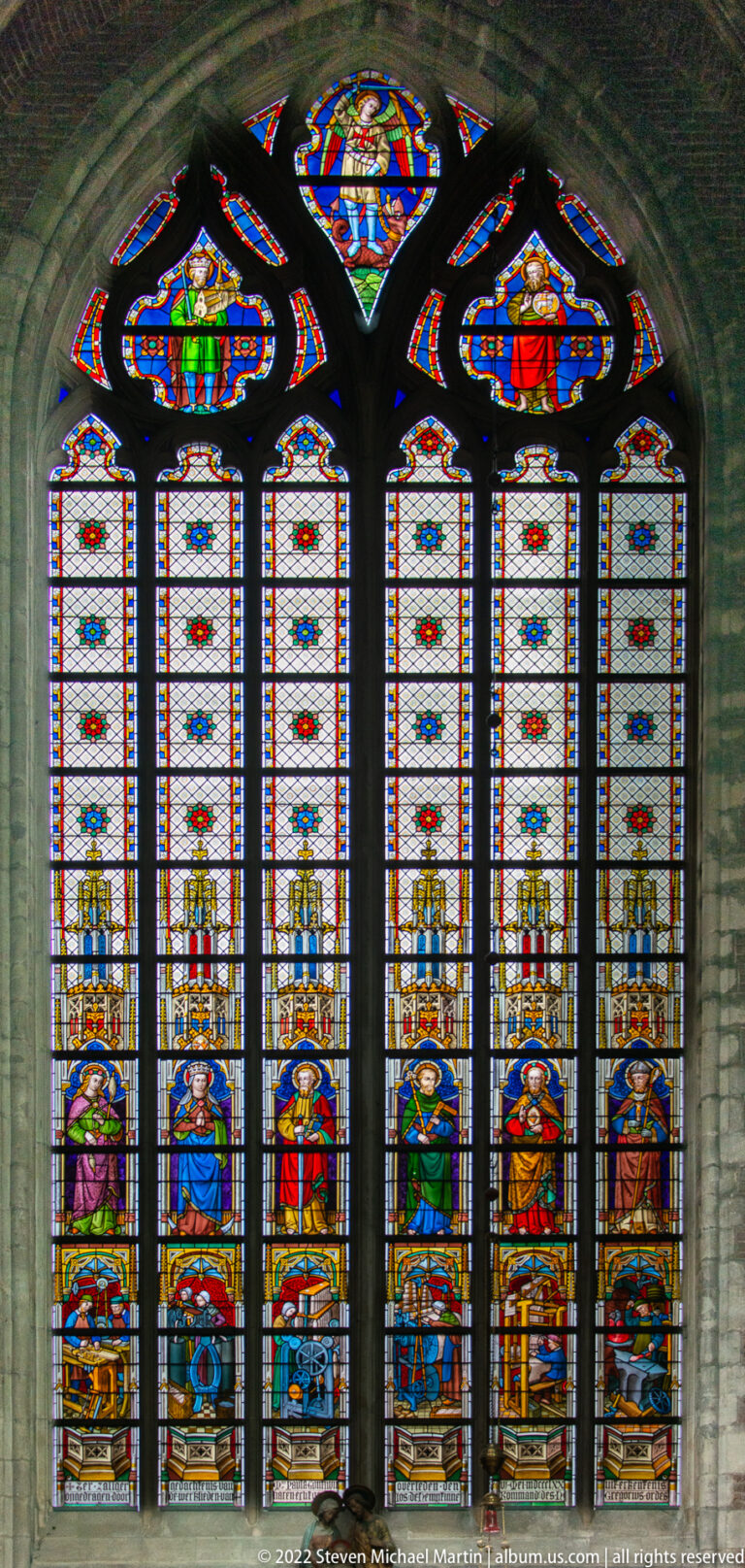 Tall Gothic stained-glass window with standing figures, heraldic shields, and colorful decorative panels.