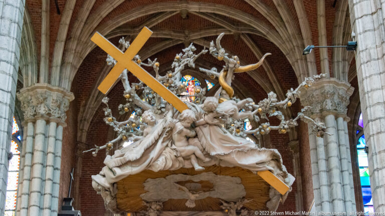 Elaborate Baroque pulpit sculpture with pale swirling figures and a large golden cross beneath cathedral vaults.