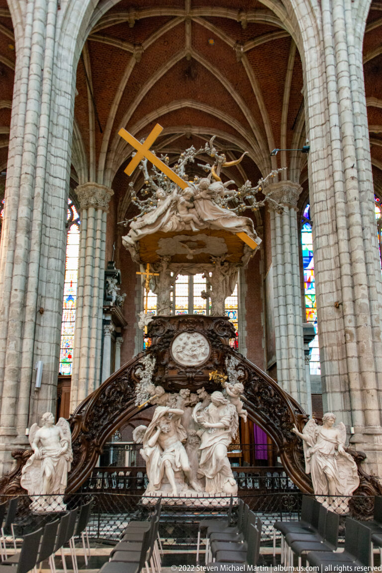Large ornate cathedral pulpit with dark carved stairs, pale sculptures, and an expansive canopy beneath ribbed vaults.
