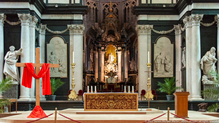 Red cloth draped over a wooden cross before an ornate cathedral altar with statues, columns, reliefs, and candles.