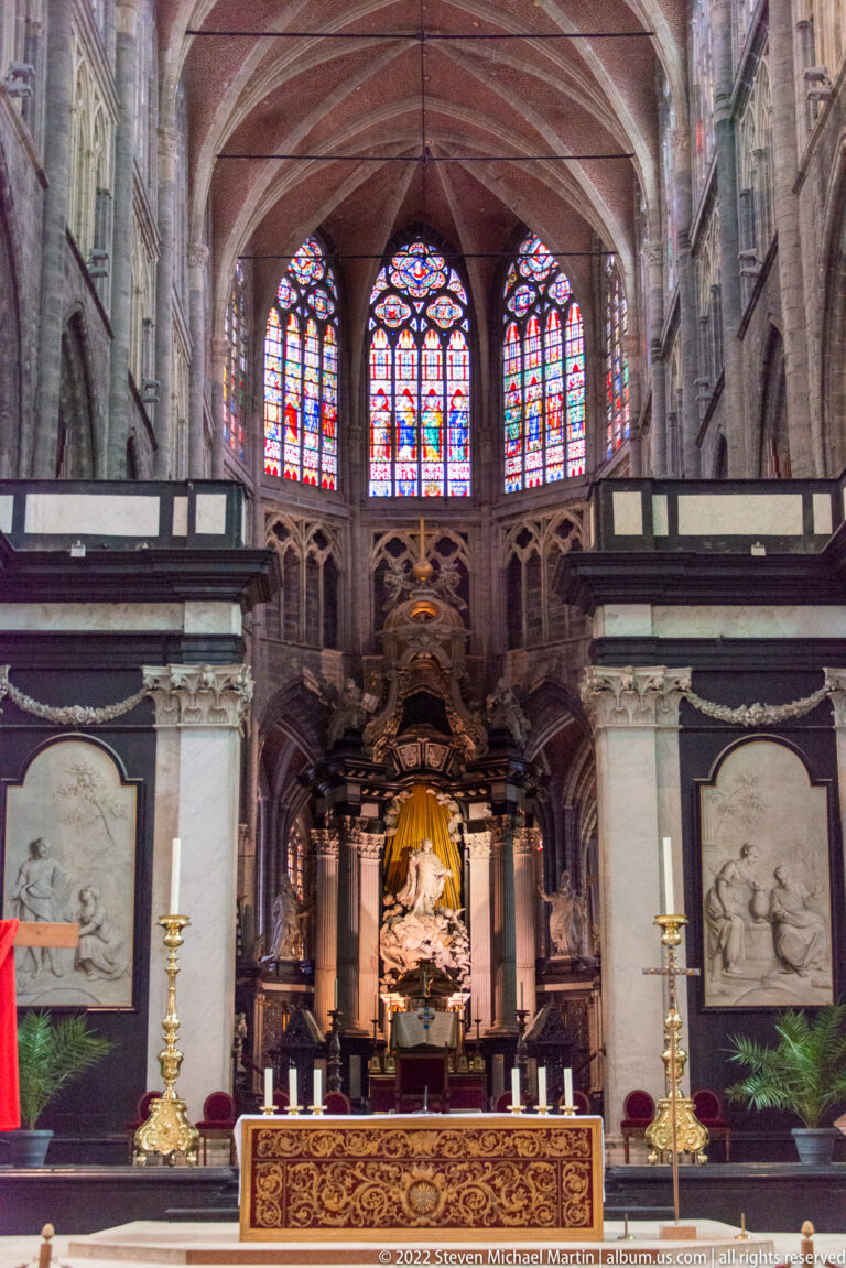 Frontal cathedral altar with dark architectural framing, pale reliefs, candlesticks, and stained-glass windows above.