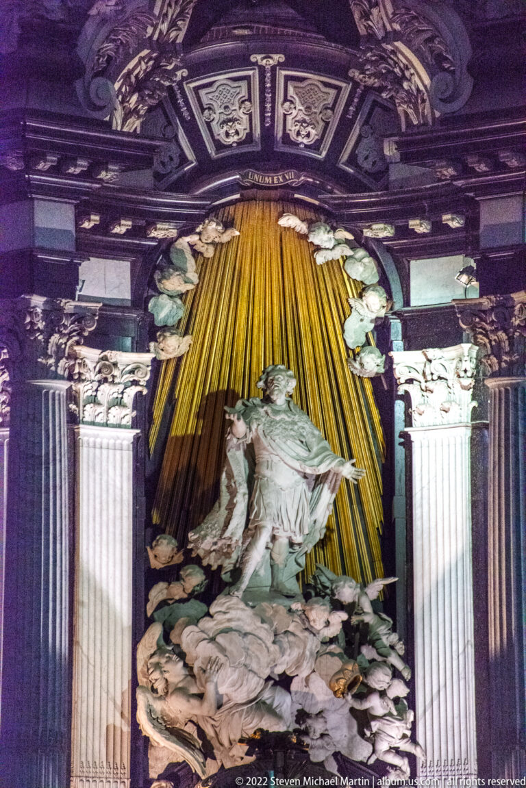 Baroque side altar with pale robed figures set against radiating gilded rays between dark framing and columns.