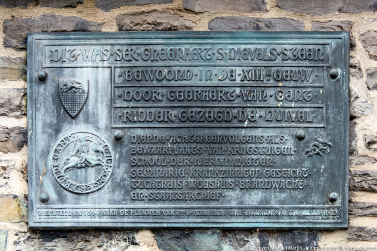 Rectangular metal commemorative plaque with raised Dutch text and heraldic emblems mounted on a stone wall.