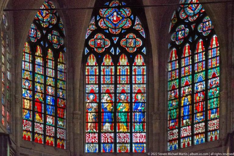 Three tall Gothic stained-glass windows filled with colorful saints, decorative panels, and pointed tracery.