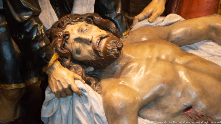Close view of the dead Christ with closed eyes, side wound, and white cloth, supported by surrounding figures.