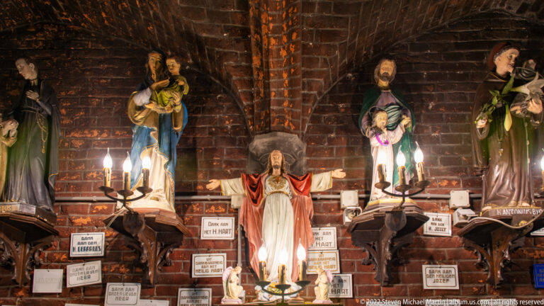Brick-walled devotional shrine with painted Marian and saint statues, candles, and numerous votive plaques.