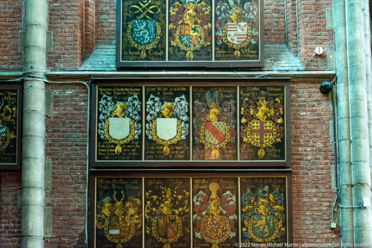 Rows of colorful heraldic shields and inscriptions mounted on a brick wall between pale stone columns.