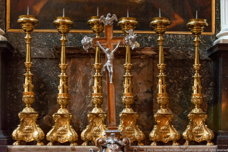 Central crucifix flanked by tall gilded altar candlesticks with ornate bases against a dark background.