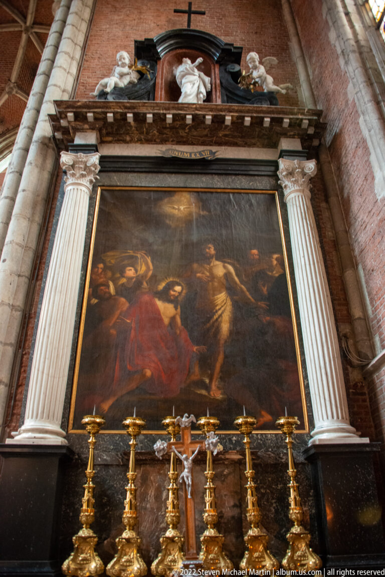 Large dark religious painting in a columned architectural frame above gilded candlesticks and a crucifix.