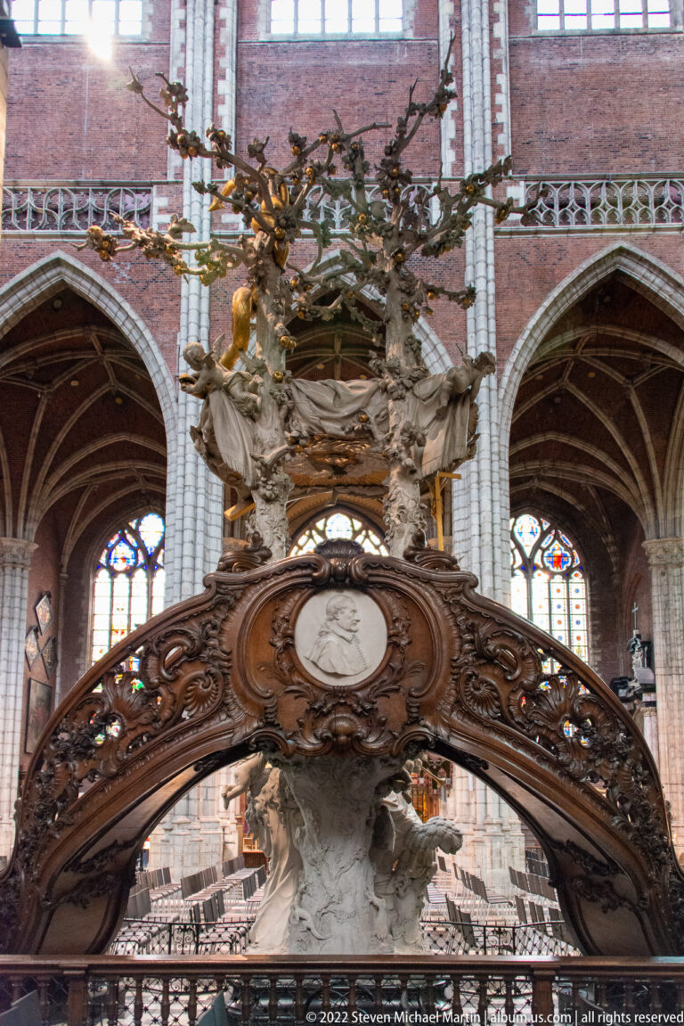 Upper portion of a monumental cathedral pulpit with dark carved wood and pale figures against Gothic arches.