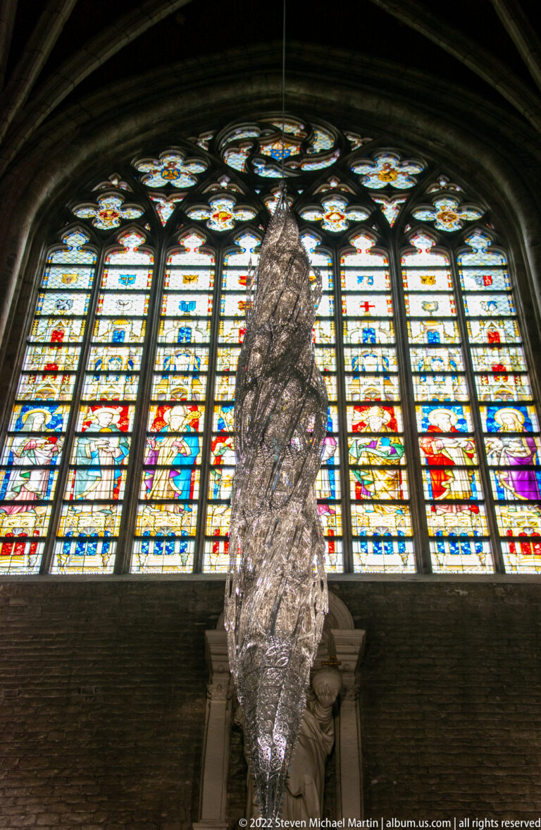 Abstract metallic hanging sculpture suspended before a large multicolored stained-glass window in a cathedral.